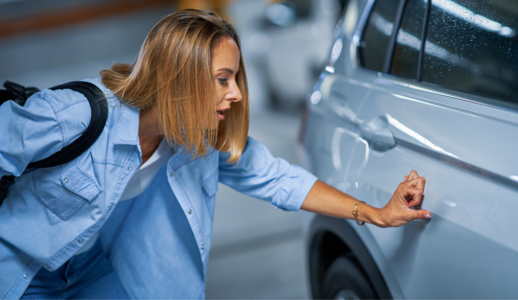 Driver examines car door scratch in parking garage, highlighting the need to fix small car dents quickly.
