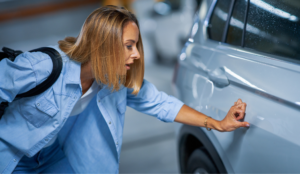 Driver examines car door scratch in parking garage, highlighting the need to fix small car dents quickly.