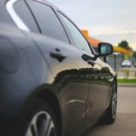 Close-up of a dark car's side, with its mirror and door visible, parked in a lot with a blurred background.