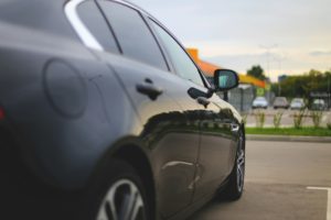 Close-up of a dark car's side, with its mirror and door visible, parked in a lot with a blurred background.