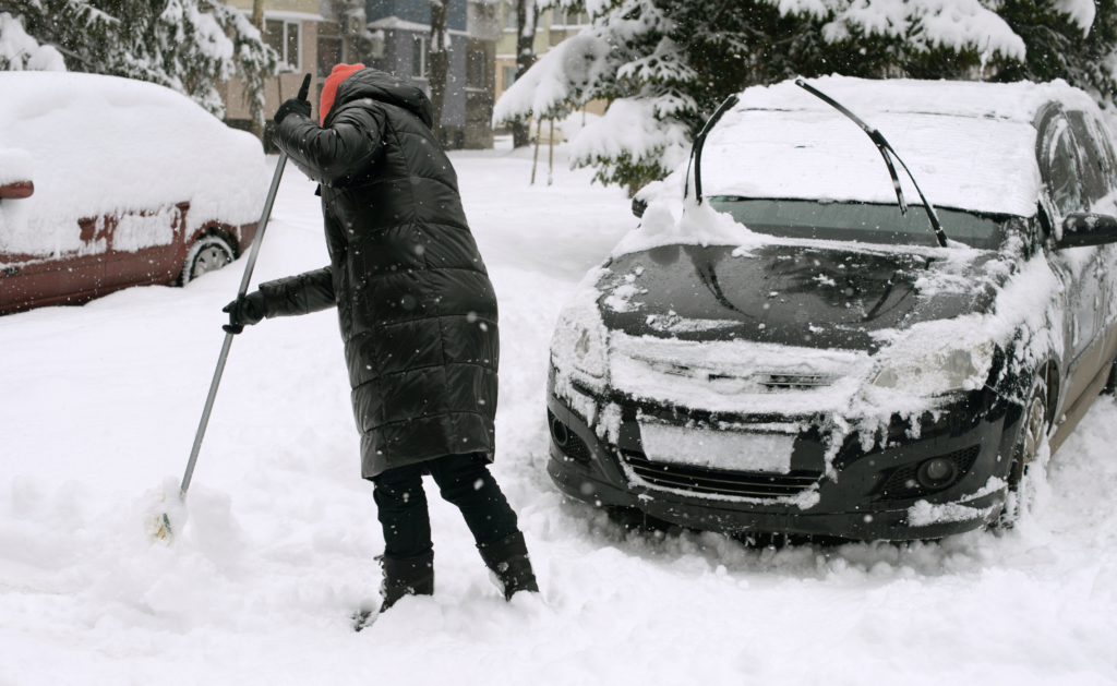 Person shoveling snow near car, showing importance of winter car care to maintain a car's exterior.