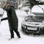 Person shoveling snow near car, showing importance of winter car care to maintain a car's exterior.