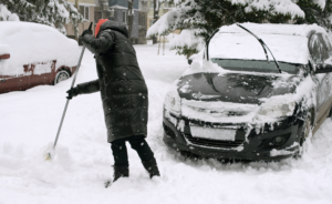 Person shoveling snow near car, showing importance of winter car care to maintain a car's exterior.